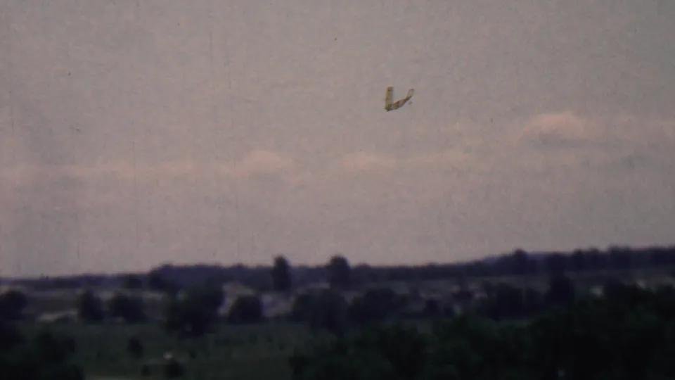 1961 home-movie frame with broad clouded sky above a low wooded horizon and distant countryside, rendered in muted 8mm tones.