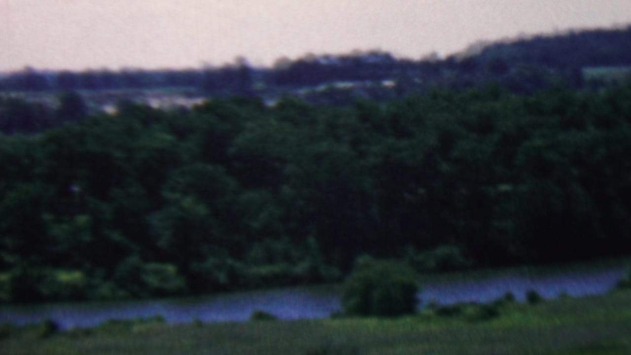 1961 home-movie frame from the Covington, Kentucky metadata showing a calm lake bordered by dense trees with rolling hills and fields in the distance.