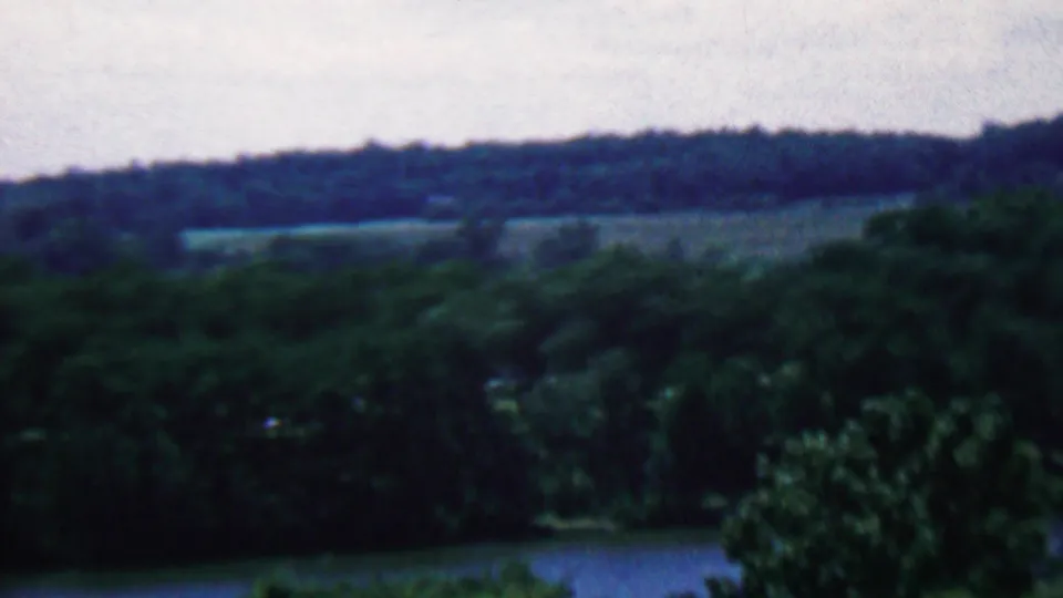 Archival 8mm frame showing a hazy view over treetops toward rolling hills and fields, with water partly visible near the lower edge.