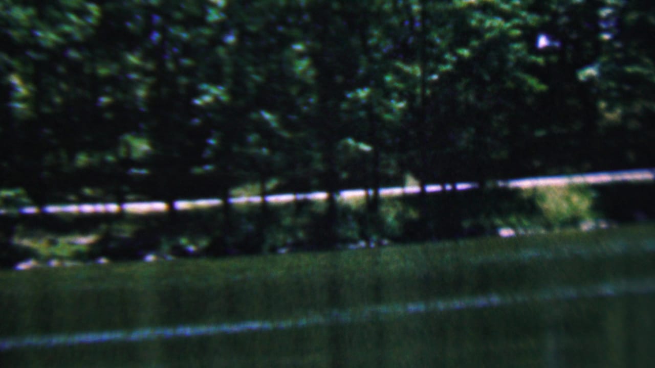 1961 Covington, Kentucky 8mm frame showing a wooded riverbank, green water, and a pale fence or rail running horizontally near the trees.