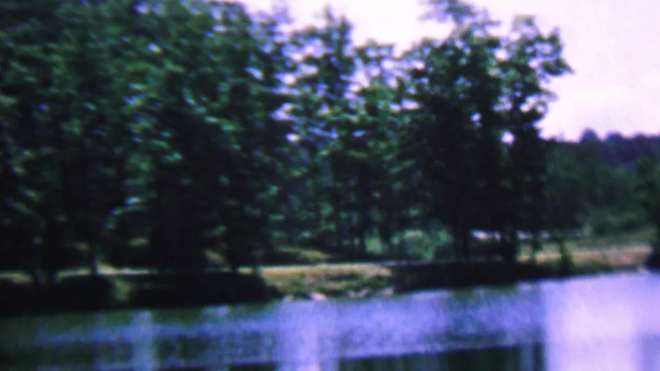 1961 8mm home-movie frame from Covington, Kentucky, showing a broad pond or lake surface beneath dense trees and a bright summer sky.