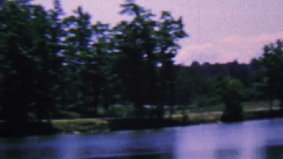 Final 1961 8mm frame showing the Covington, Kentucky lakeside scene with calm water, darkened trees, shoreline vegetation, and a pale sky.