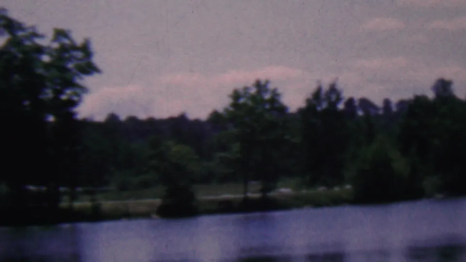 A handheld 8mm view continues across the lakeside trees and far bank, with muted 1960s color and soft focus.