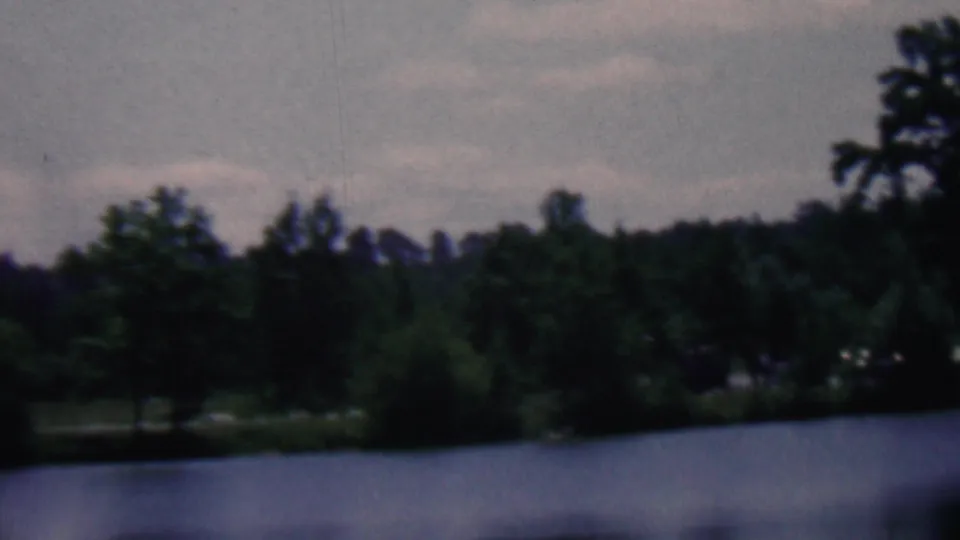 The camera pans along a wooded pond or lake edge in the metadata-supported Kentucky camping footage, with shrubs and a small pale distant shape partly hidden by trees.