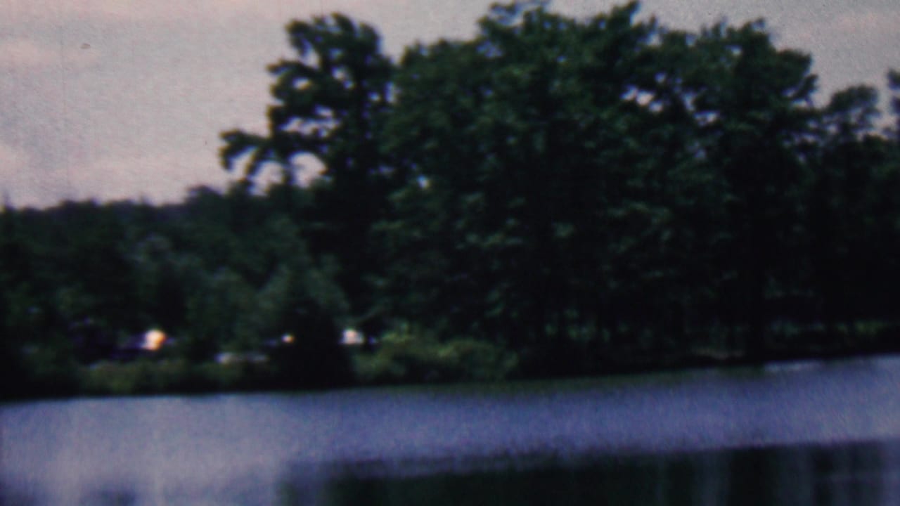 1961 home-movie frame associated with Covington, Kentucky showing a calm lake, wooded shoreline, and soft cloudy sky with muted 8mm color.