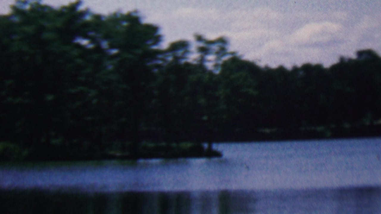 1961 home-movie frame associated with Covington, Kentucky showing a wooded lakeside point, dark forest background, and still water beneath an overcast sky.