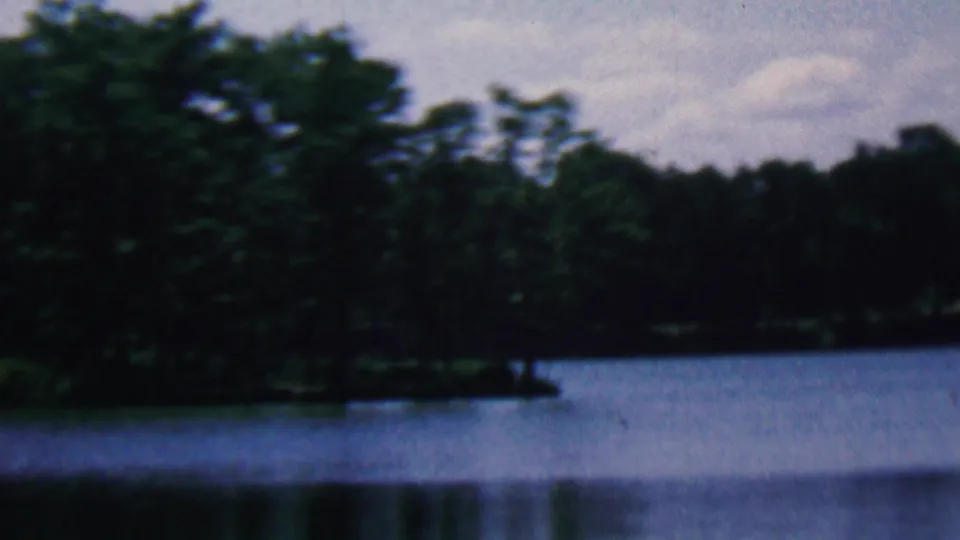 1961 home-movie frame associated with Covington, Kentucky showing a wooded lakeside point, dark forest background, and still water beneath an overcast sky.