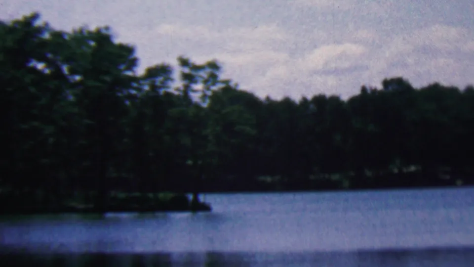 1961 home-movie frame associated with Covington, Kentucky showing a blue-toned lake, dense shoreline trees, and a softly blurred forest edge.