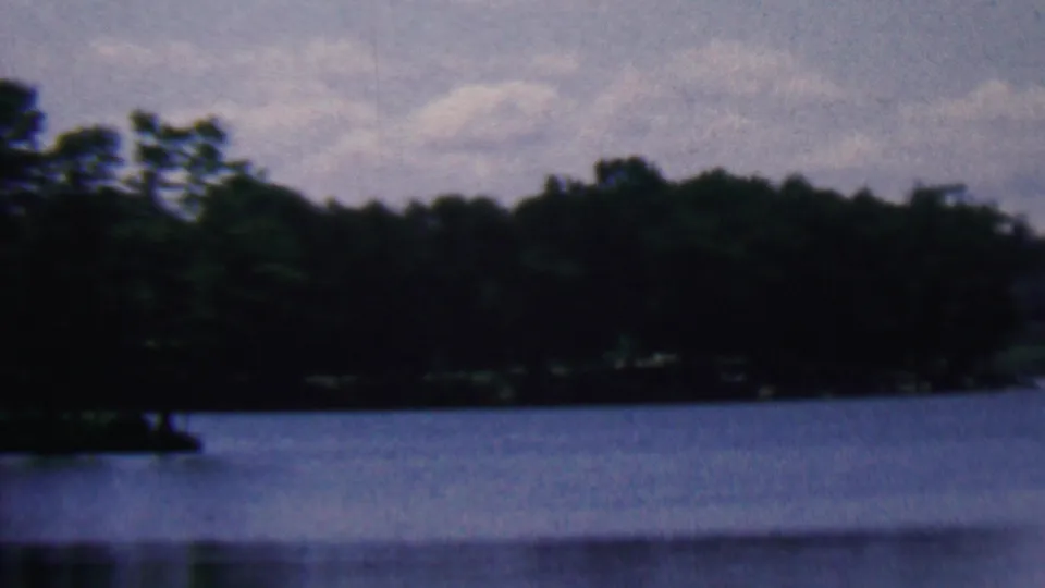 1961 home-movie frame associated with Covington, Kentucky showing calm lake water, a shadowed tree line, and a pale clouded sky in soft 8mm color.