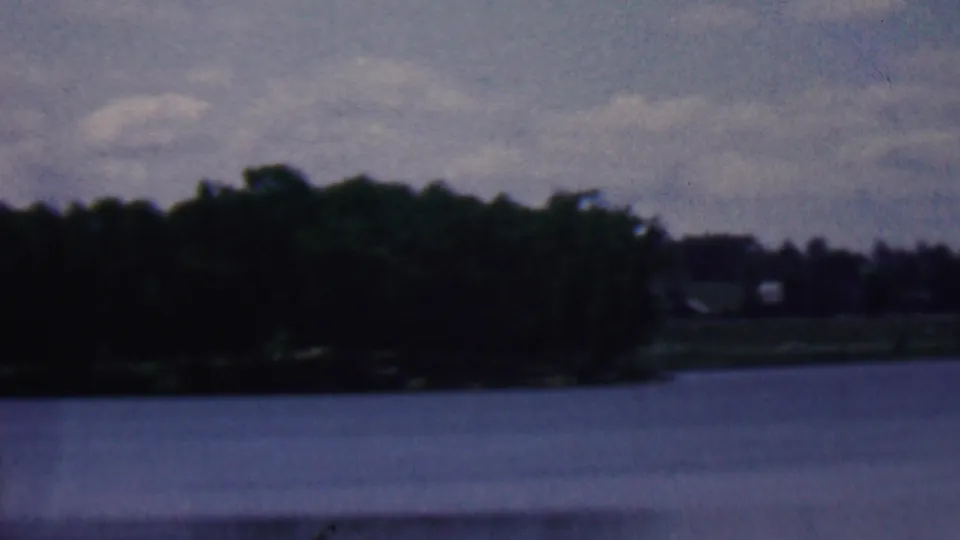 1961 8mm home-movie frame from Covington, Kentucky metadata with open water, a wooded far shore, and indistinct distant structures beneath a cloudy sky.