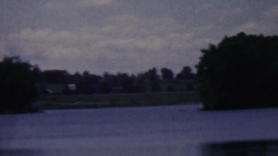 1961 8mm home-movie frame from Covington, Kentucky metadata showing lake water, a dark tree mass on the right, and a low distant shoreline under clouds.