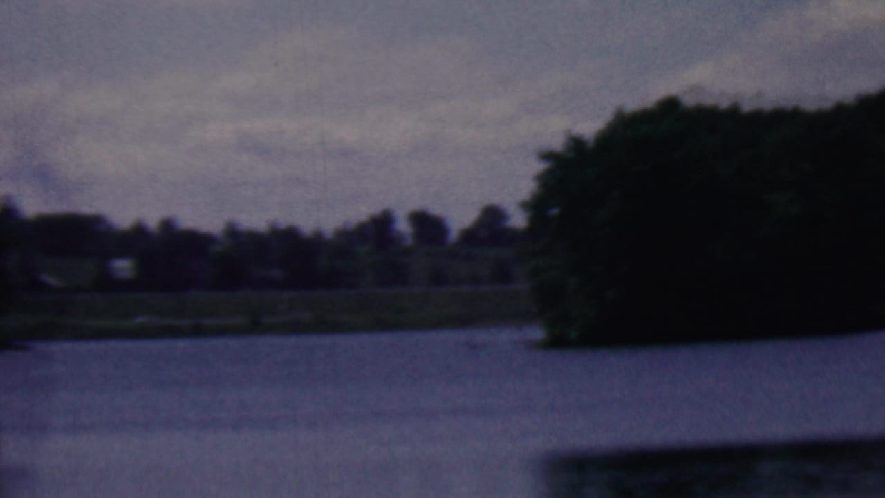 1961 Covington, Kentucky 8mm frame showing calm lake water, a distant wooded shoreline, and low gray clouds with soft film grain.