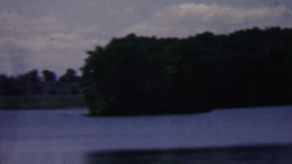 1961 Covington, Kentucky 8mm frame with dark trees along the lake edge, muted water, and an overcast blue-gray sky.