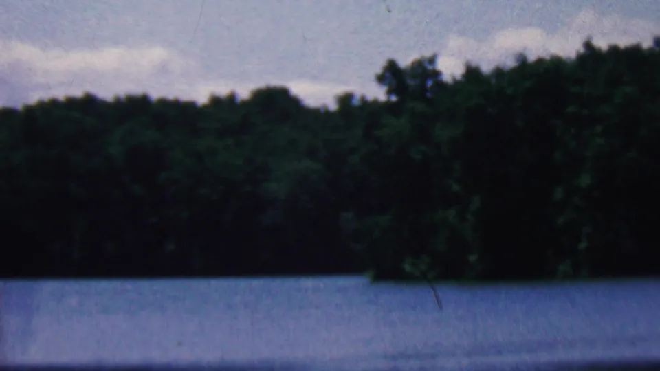 1961 Covington, Kentucky 8mm frame showing the lake foreground, a wooded shoreline, and low gray clouds during a slow scenic pan.
