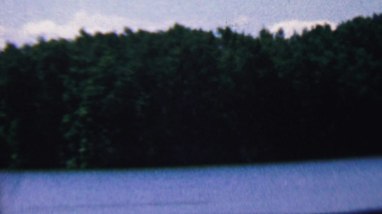 1961 Covington, Kentucky home-movie frame showing a calm blue lake approaching a dense, dark forested shoreline under a pale sky.