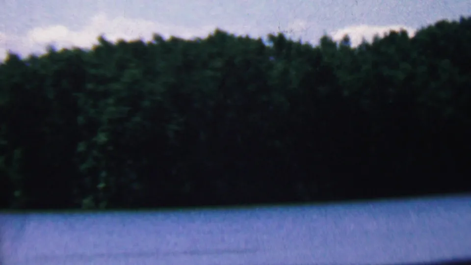 1961 Covington, Kentucky home-movie frame showing a calm blue lake approaching a dense, dark forested shoreline under a pale sky.