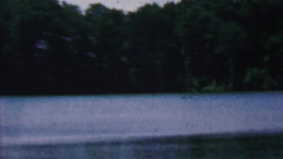1961 8mm home-movie frame showing a wooded Kentucky lakeshore, blue water, and a narrow darker foreground edge near the camera.