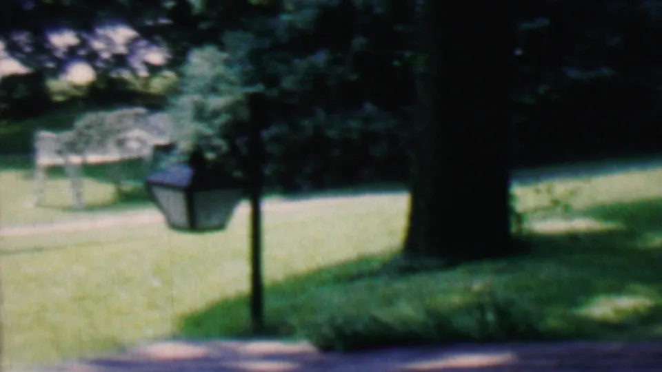 1956 Hatfield, Pennsylvania home-movie frame with a dark lamppost beside a path, a grassy yard, mature trees, and soft-focus landscaping in the background.