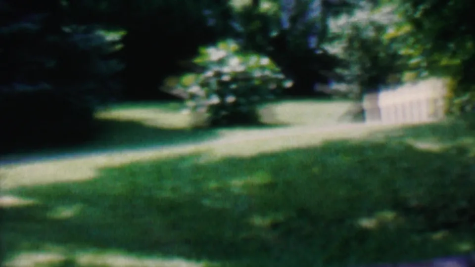 1956 Hatfield, Pennsylvania home-movie frame looking across a green backyard toward a pale fence, shaded trees, shrubs, and a curving light-toned path or lawn edge.