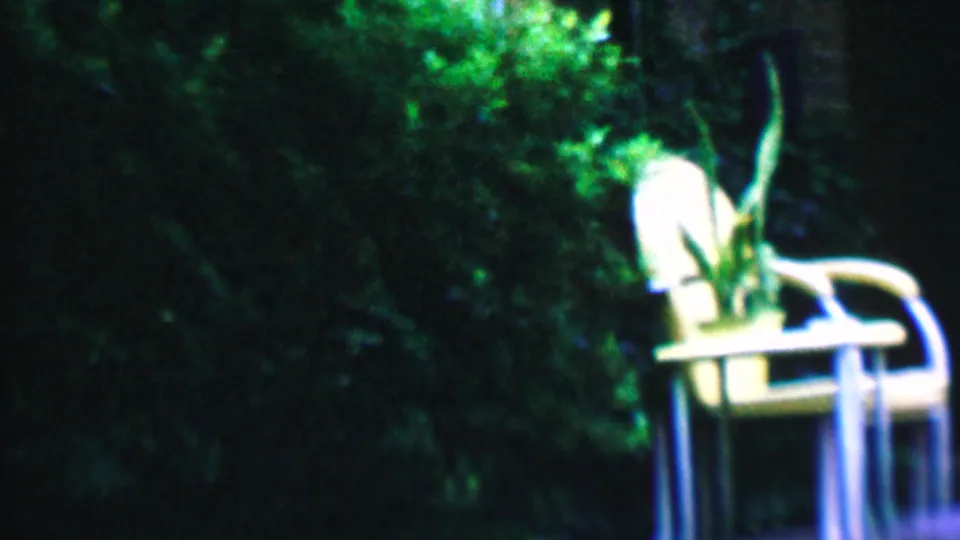 A white outdoor chair holding or standing beside a potted plant appears against dark backyard foliage in the 1956 Hatfield home-movie scene.