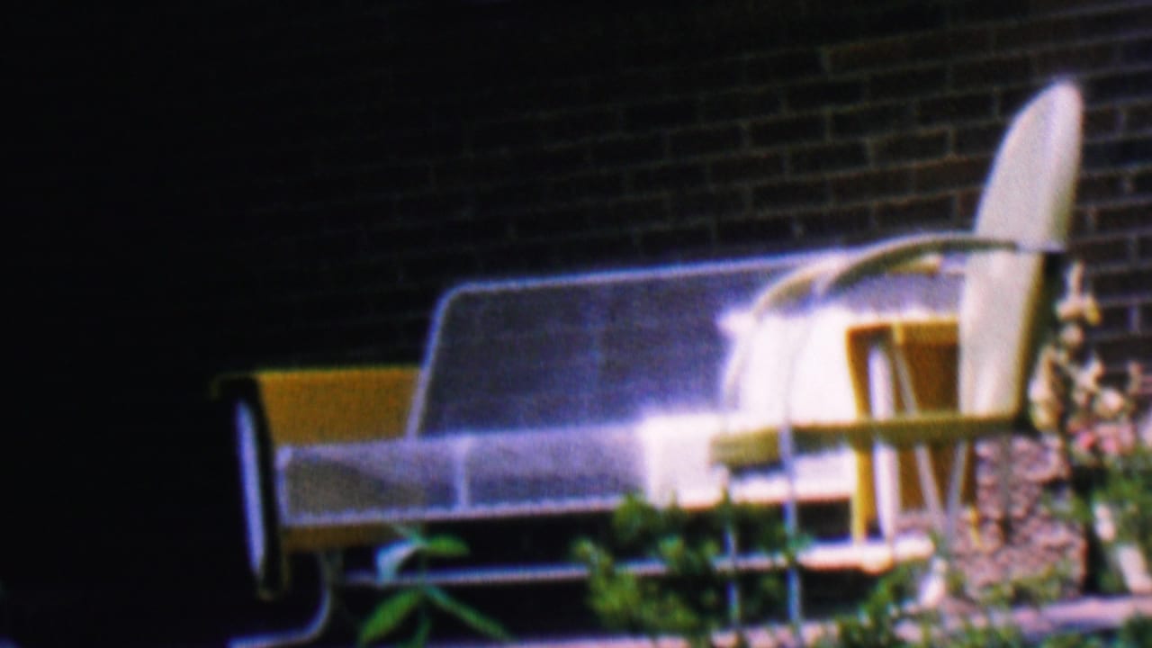 1956 Hatfield, Pennsylvania home-movie frame showing yellow and white metal patio seating near a red brick house exterior with plants in front.
