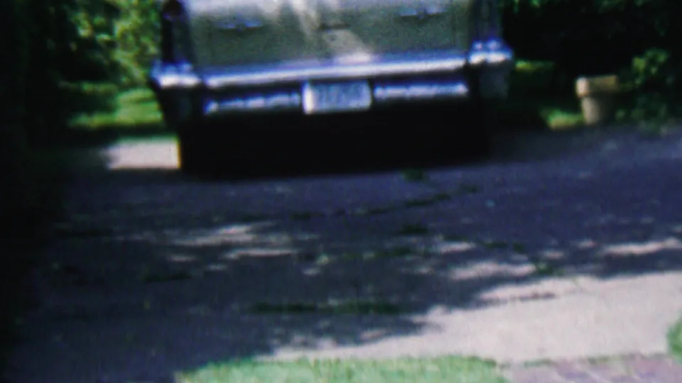 1956 Hatfield, Pennsylvania frame showing the rear of a dark mid-century automobile parked on a driveway beside a brick house and yard plants.