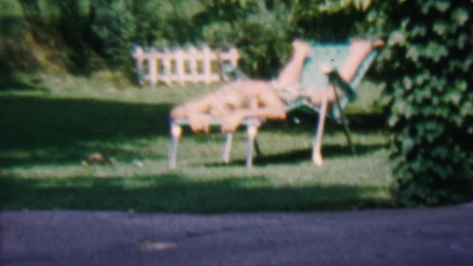 1956 Hatfield, Pennsylvania 8mm frame showing a person sunbathing facedown on a lounge chair near ivy, grass, and a white fence.