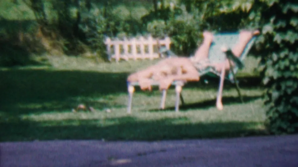 1956 Hatfield, Pennsylvania 8mm frame with a person lying facedown on a backyard lounge chair beside ivy, lawn, and a white picket fence.