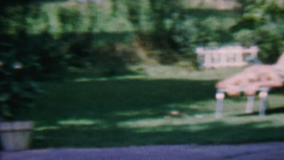 A 1956 Hatfield, Pennsylvania garden frame shows lawn furniture, white fencing, trees, a potted plant, and strong sunlight across the grass.