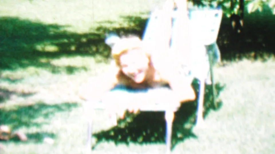 A young white woman smiles from a lawn chair in a bright 1956 Hatfield, Pennsylvania garden scene, with green grass and foliage around the sunlit seating area.