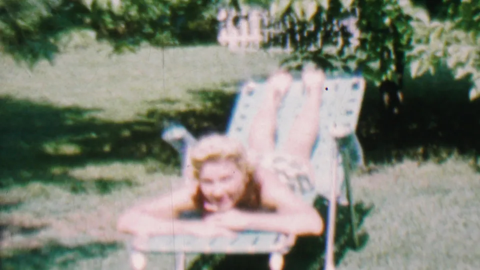 The 1956 home-movie frame shows the young woman propped on her elbows on a metal lawn chair, looking toward the camera in a shaded grassy backyard.