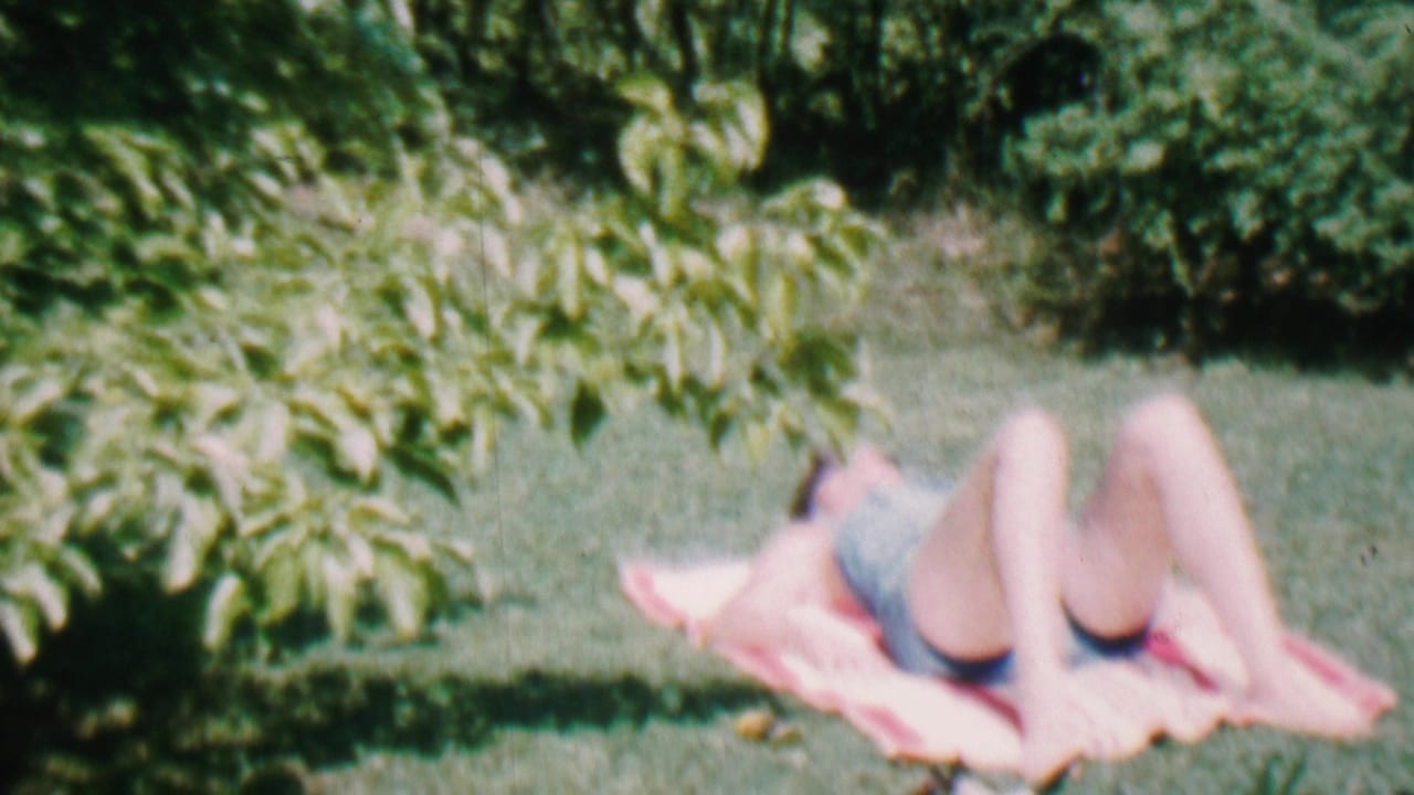 Person lying on a pink towel on grass with shrubs behind them.