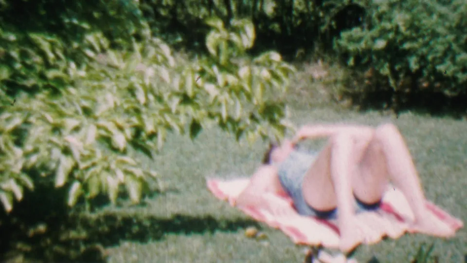 The frame shows a relaxed figure lying on a pink towel on grass under overhanging leaves, in archival 8mm footage identified as Hatfield, Pennsylvania in 1956.
