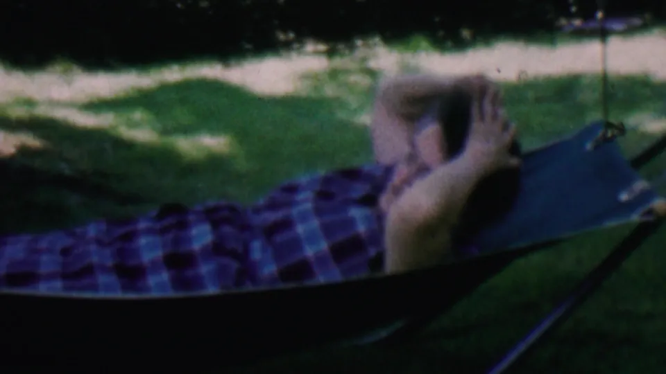 A man in a blue plaid shirt reclines in a dark hammock on a shaded lawn, with greenery and a bright path or open ground beyond.