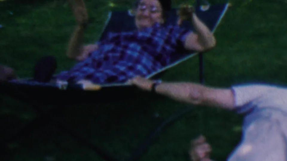 The older man appears to laugh or gesture while sitting partly upright in the hammock, with a partial nearby person visible at the edge of the 1956 home-movie frame.