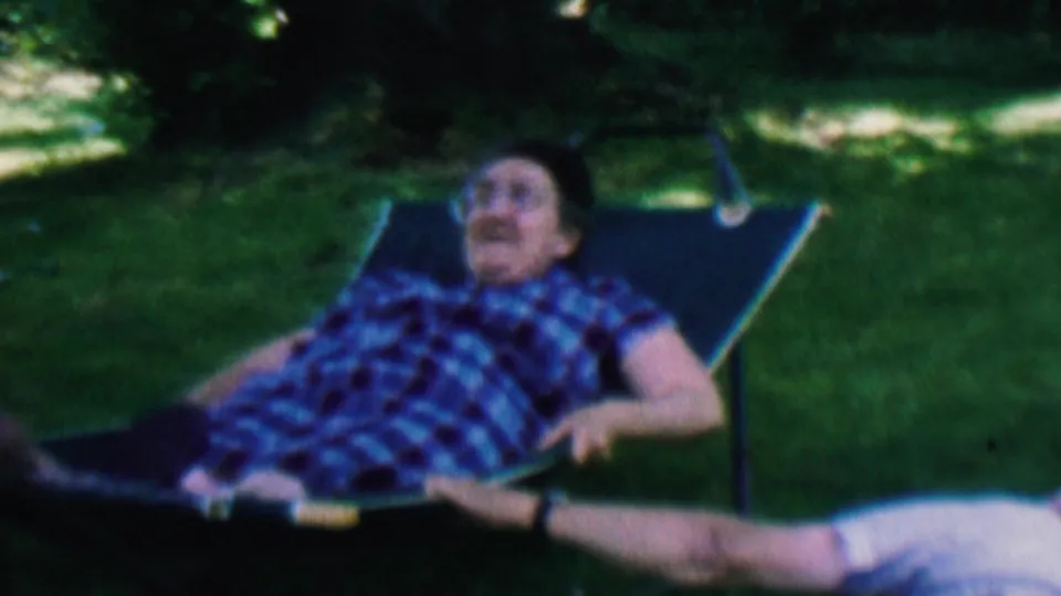 A smiling man reclines in a backyard lawn chair in Hatfield, Pennsylvania, with another adult partially visible lying on the grass nearby.
