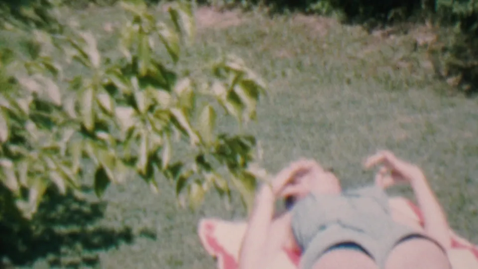 An adult lies on a blanket on the grass under leafy branches during a sunny backyard moment in the 1956 Hatfield home movie.