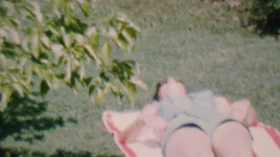 An adult in a bathing suit lies back on a blanket in the grassy backyard, filmed from a slightly elevated home-movie angle.