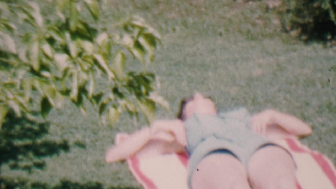 In Hatfield, Pennsylvania, 1956 home-movie footage, a woman in a light swimsuit lies on a red and white striped towel on a sunlit grassy lawn.