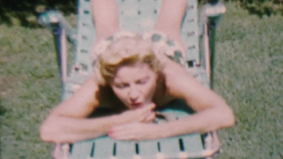An adult woman in a patterned swimsuit lies on a green lawn chair in a grassy Hatfield, Pennsylvania backyard and looks toward the camera in this 1956 8mm frame.
