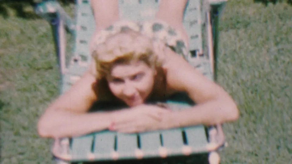 The woman smiles at the camera while lying on her stomach on a green lawn chair, with grass and bright outdoor light visible in this 1956 Hatfield home-movie frame.