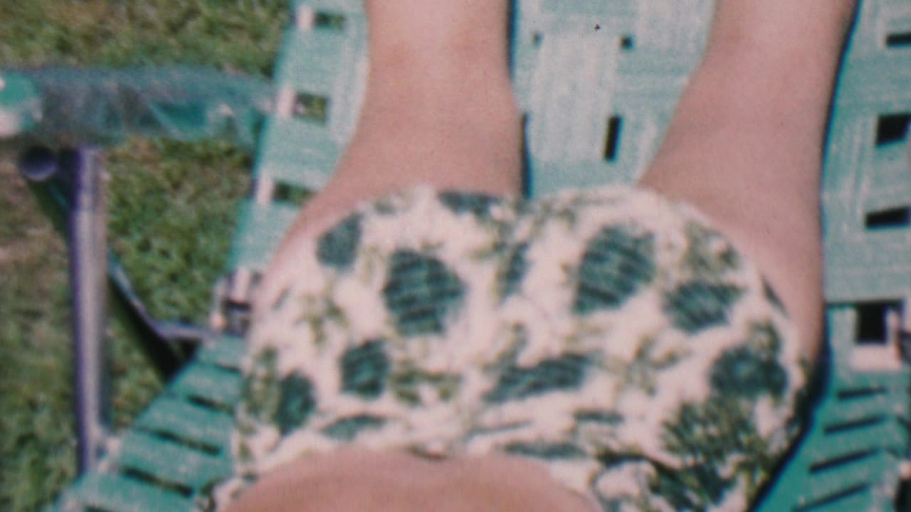 1956 Hatfield, Pennsylvania home-movie frame showing a person reclining outdoors in a white and green leaf-print swimsuit on a green lawn chair.