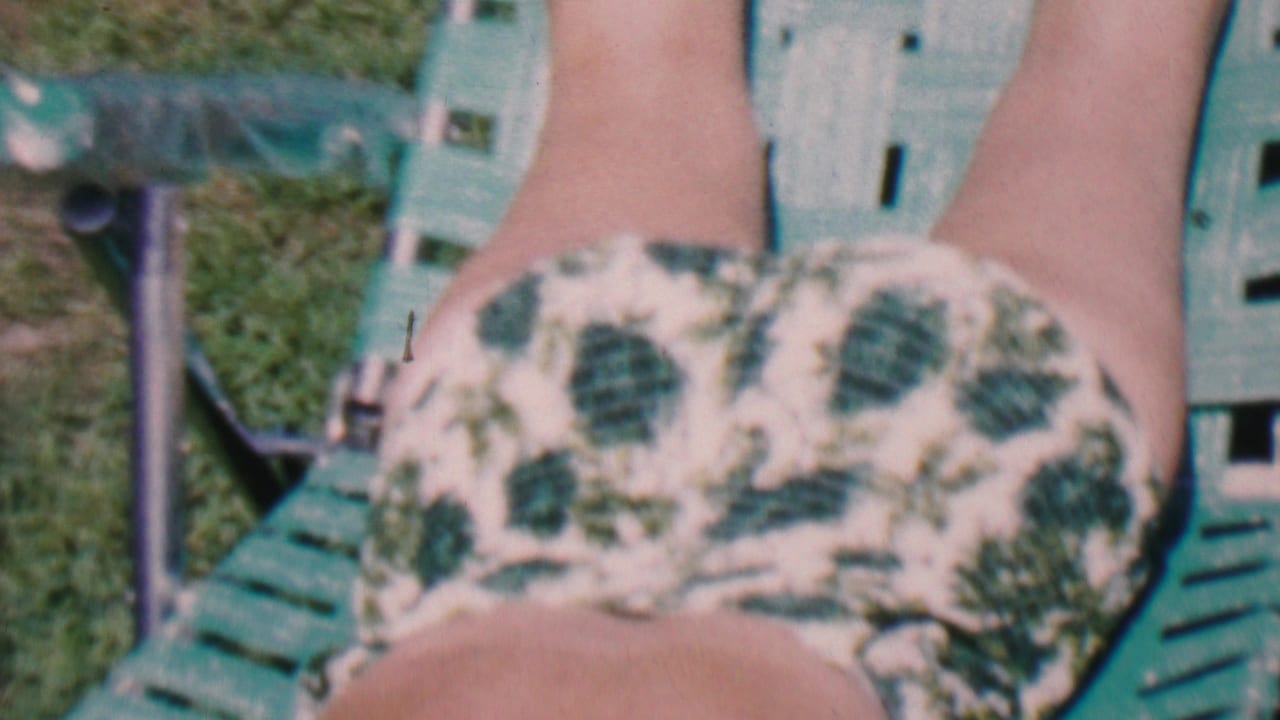 Close 1956 home-movie view of a person’s lower body in a white and green leaf-print swimsuit while reclining on a green lawn chair outdoors.