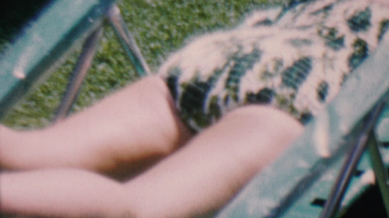 1956 Hatfield, Pennsylvania home-movie frame showing a person sunbathing on a lawn chair in a patterned swimsuit, partly obscured by the chair frame.