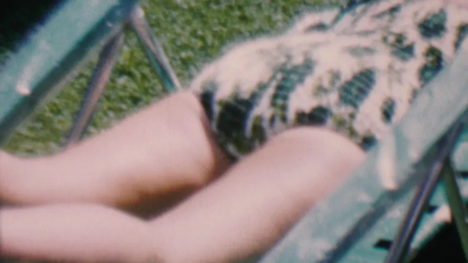 1956 Hatfield, Pennsylvania home-movie frame showing a person sunbathing on a lawn chair in a patterned swimsuit, partly obscured by the chair frame.