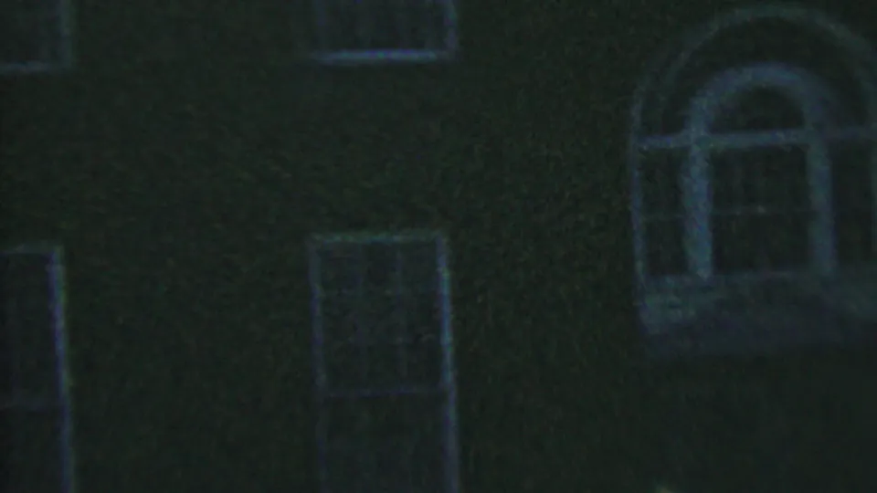 Low-light 1956 8mm frame from Hatfield, Pennsylvania showing a dark brick building facade with rectangular window openings and pale trim.