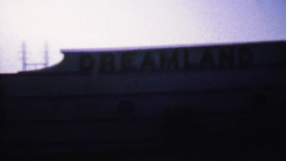Wider dusk frame showing the Dreamland lettering along a low roofline, with faint utility structures silhouetted in the background.
