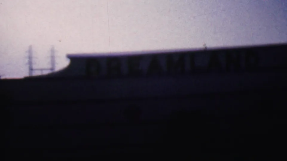 Late-frame 8mm view of the Dreamland sign partially darkened by low exposure, with the roofline and pale sky still visible.
