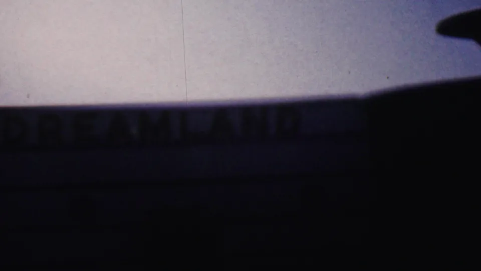 The camera continues past the shadowed DREAMLAND facade, with the sign softened by motion blur and the sky filling the upper background.