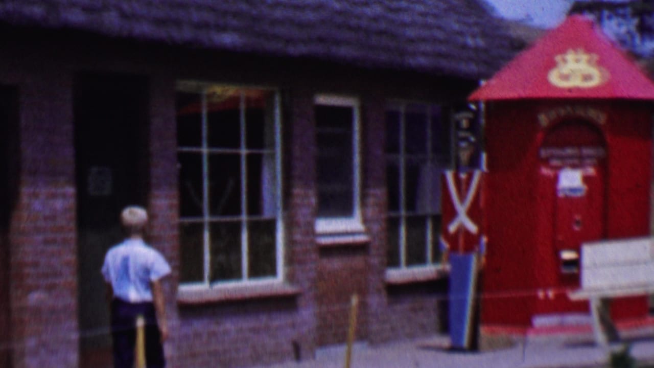 1955 Solvang street view with a male figure approaching a red post-office-style kiosk, brick storefront, white-paned windows, and a toy soldier figure.